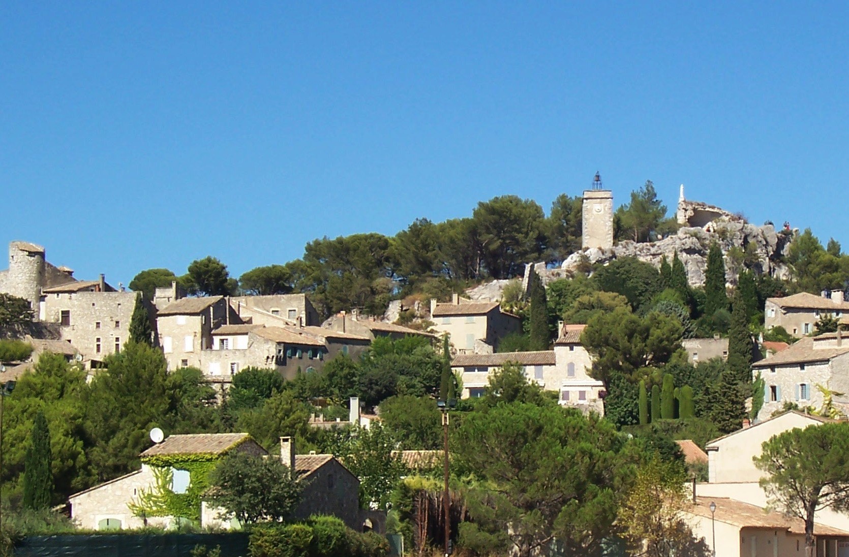 A scenic view of a hilltop village with stone buildings and a prominent tower under a clear blue sky, surrounded by lush greenery.